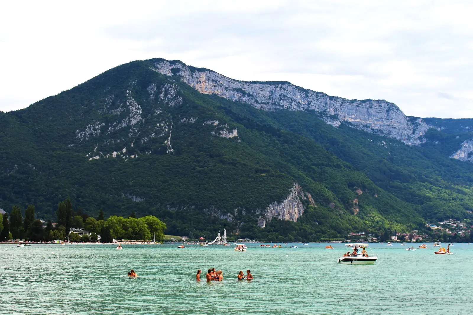 Baigneurs et pédalo sur l'eau turquoise d'un lac, grande montagne en arrière-plan