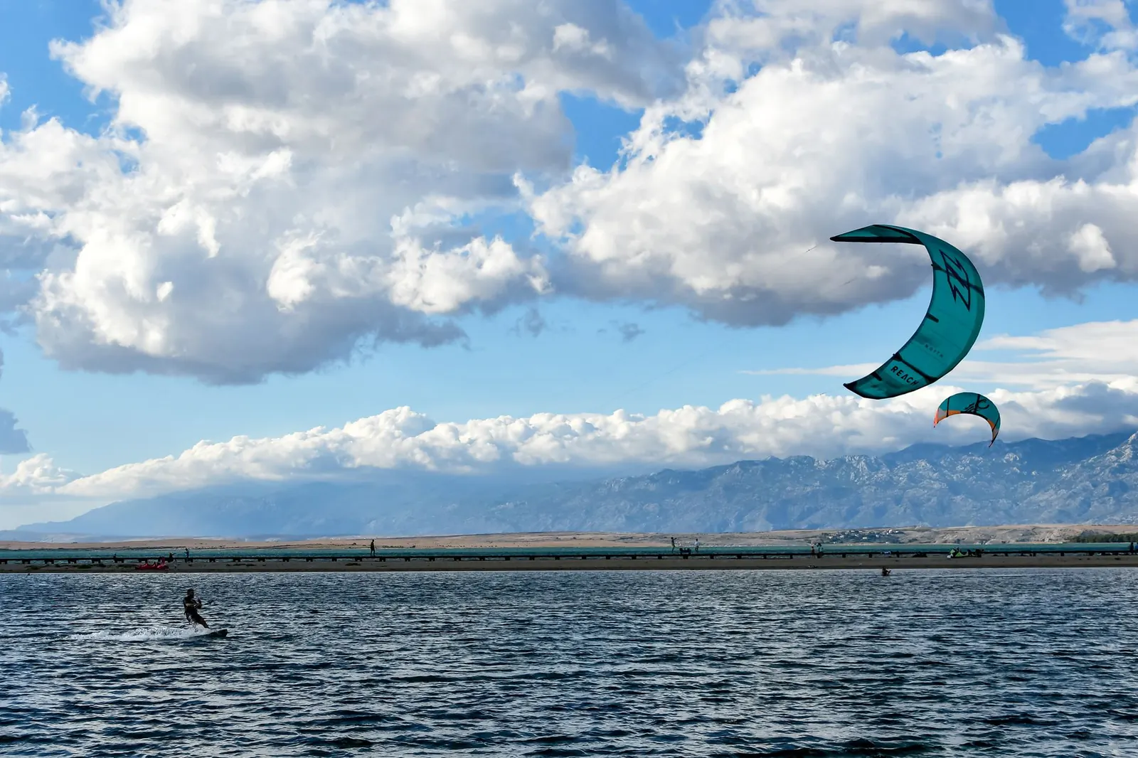 Aile de kitesurf turquoise dans un ciel nuageux au-dessus d'un lac, montagnes au loin